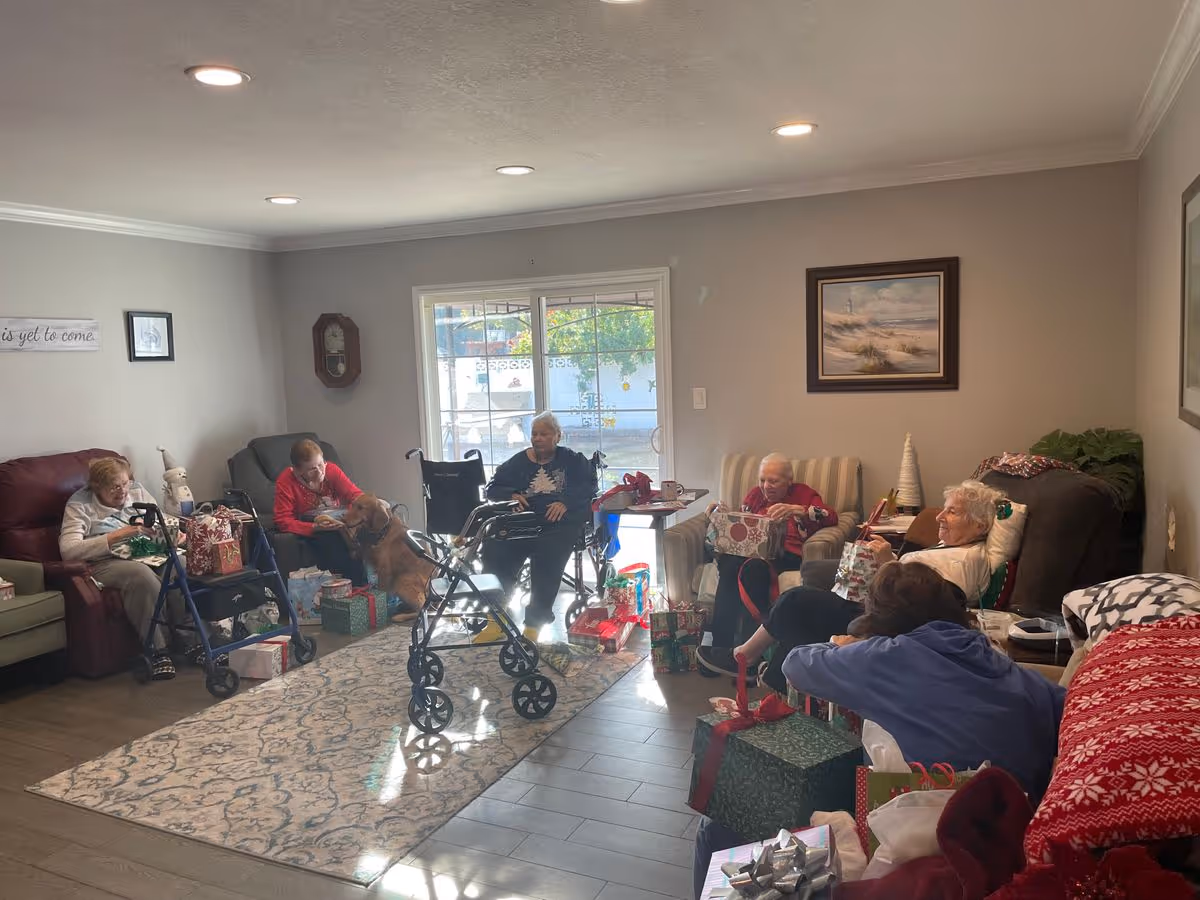 Several elderly residents sit in a living-room common area with walkers, presents, and a dog near a sliding glass door.