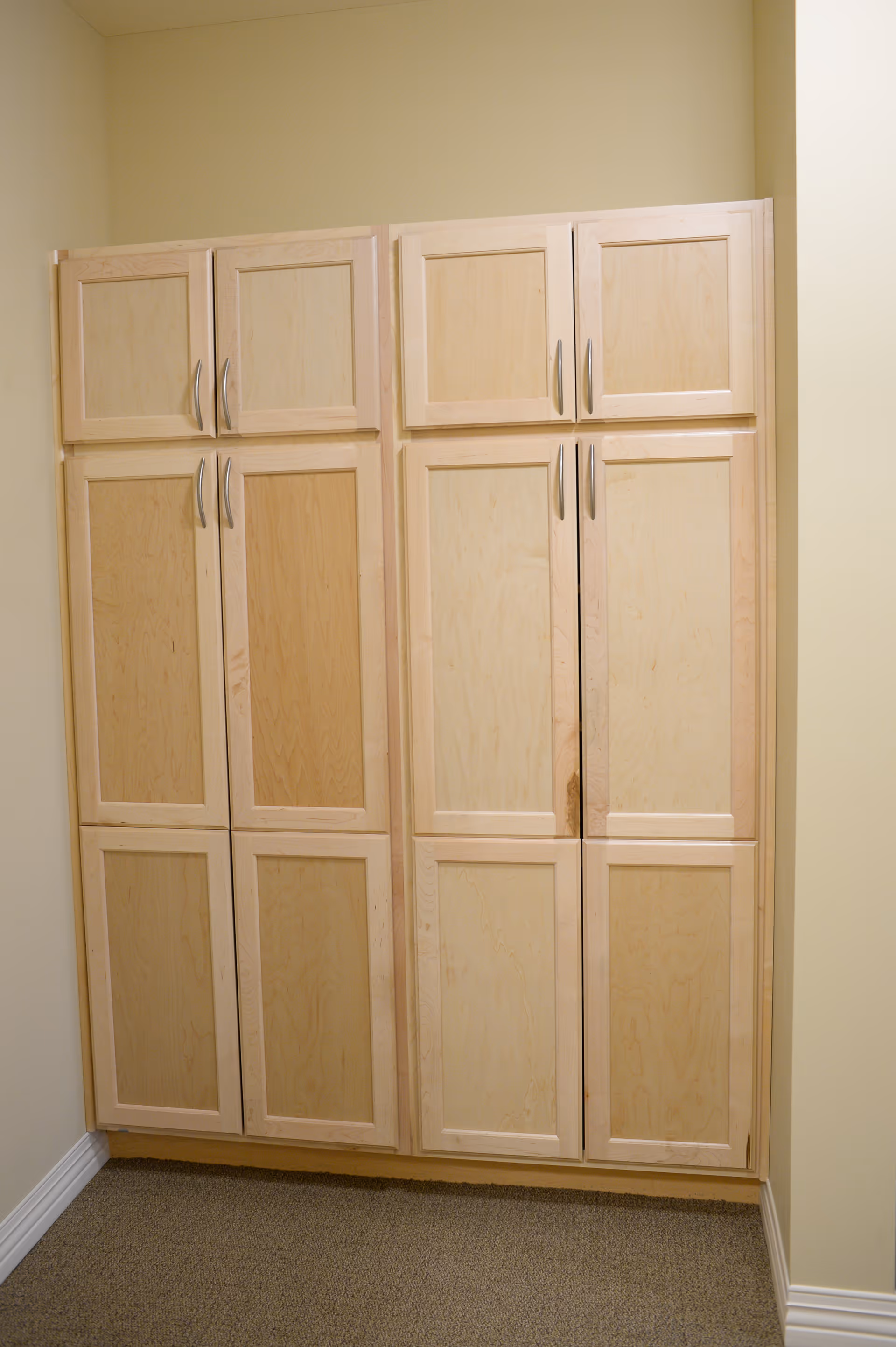A set of light wood cabinets with multiple doors and silver handles against a beige wall and carpeted floor.