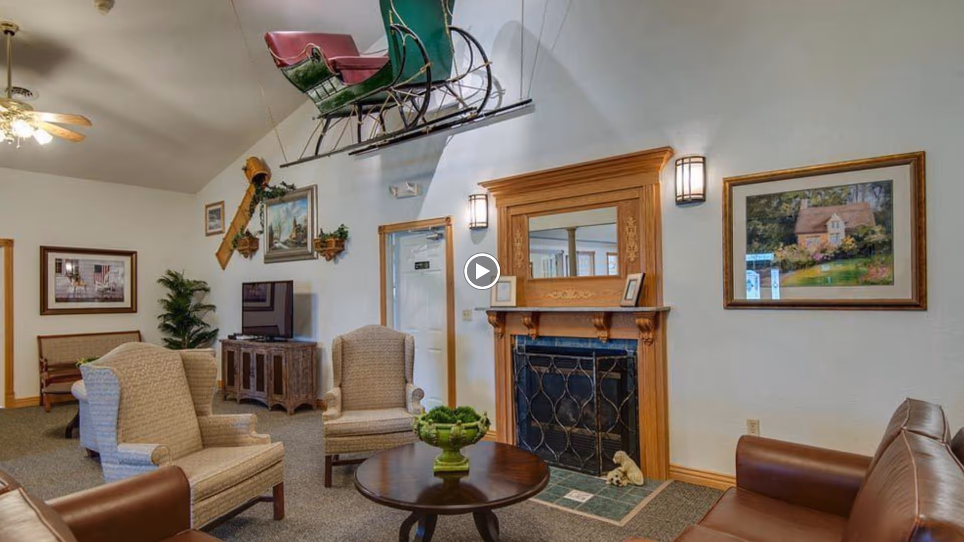 A cozy lounge with upholstered armchairs around a round coffee table, a decorative fireplace with wooden mantel, TV cabinet, framed artwork, and a sleigh suspended from the ceiling.