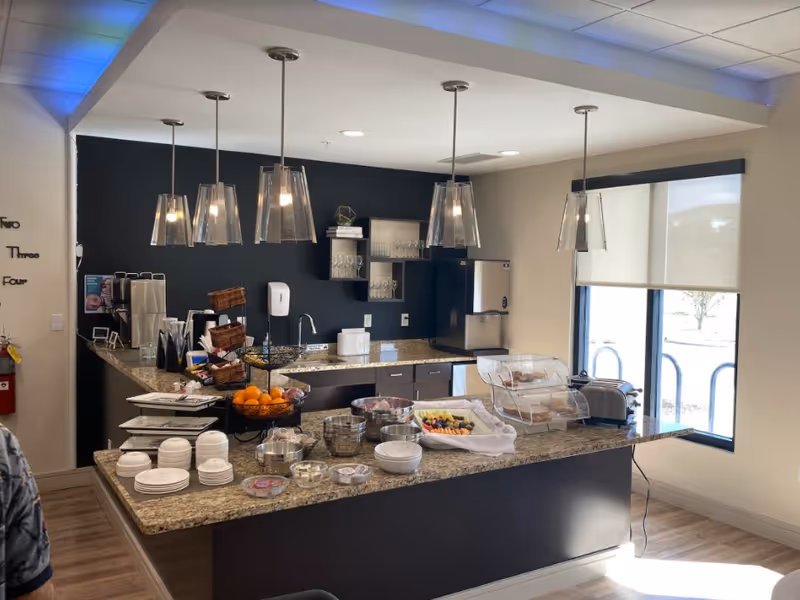 Kitchenette and buffet counter with pendant lights and a granite island stocked with plates, fruit, and serving dishes.