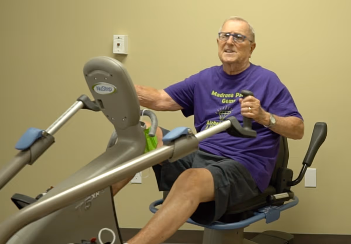 An elderly man wearing glasses and a purple t-shirt is exercising on a recumbent stationary bike in a room with beige walls.