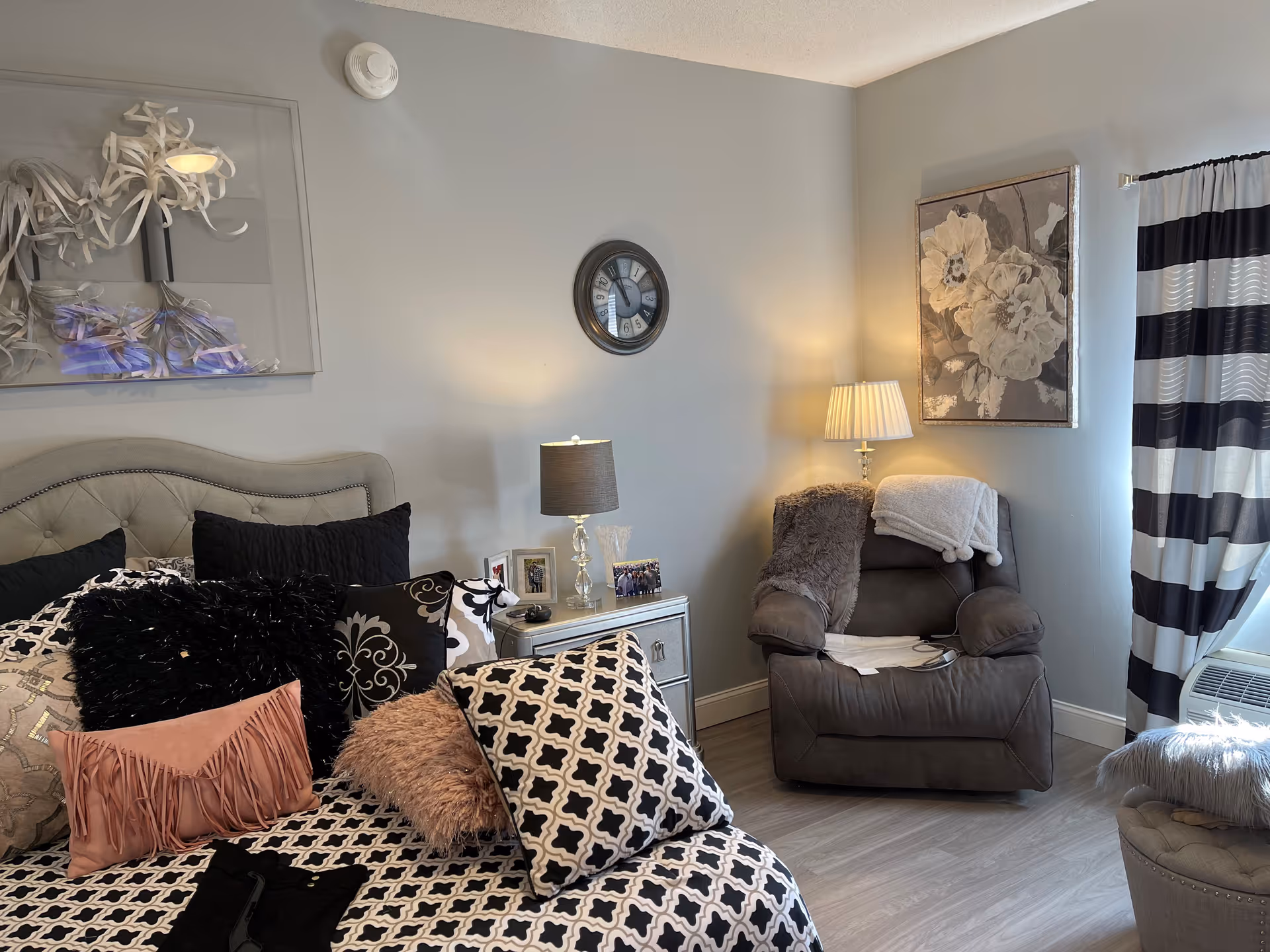 A cozy bedroom with a tufted headboard bed adorned with multiple decorative pillows in black, white, and pink tones. A bedside table with a lamp, photo frames, and a clock on the wall above. To the right, a comfortable armchair with a throw blanket and a lamp beside it. The room has light gray walls, a floral painting, striped curtains, and wood flooring.