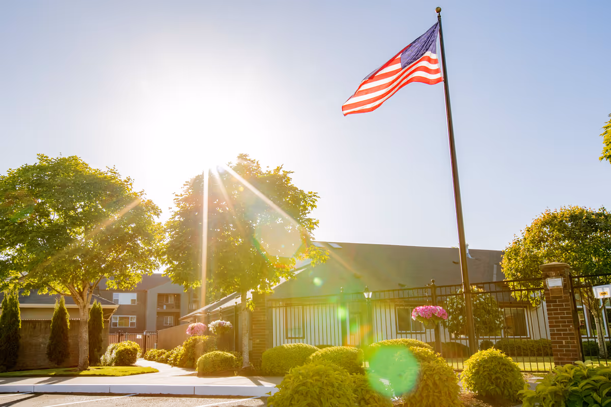 Sunlit outdoor view of a senior living facility with an American flag on a tall flagpole, green trees, bushes, and a black metal fence surrounding the property.
