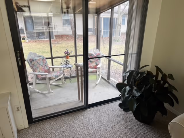 Interior view through a sliding glass door to a screened porch with two floral-cushioned rocking chairs and a small side table, with a potted plant inside.