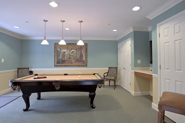 A well-lit game room with a pool table in the center, three pendant lights hanging above it, a framed painting on the light blue wall, two chairs placed against the walls, and a small counter area with a stool on the right side.