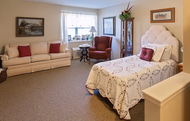 A cozy bedroom in a senior living facility featuring a single bed with a white quilted bedspread and red accent pillows, a beige sofa with red cushions, a red armchair, a small round table with a lamp, a window with sheer curtains, framed artwork on the walls, and a glass display cabinet with flowers on top.