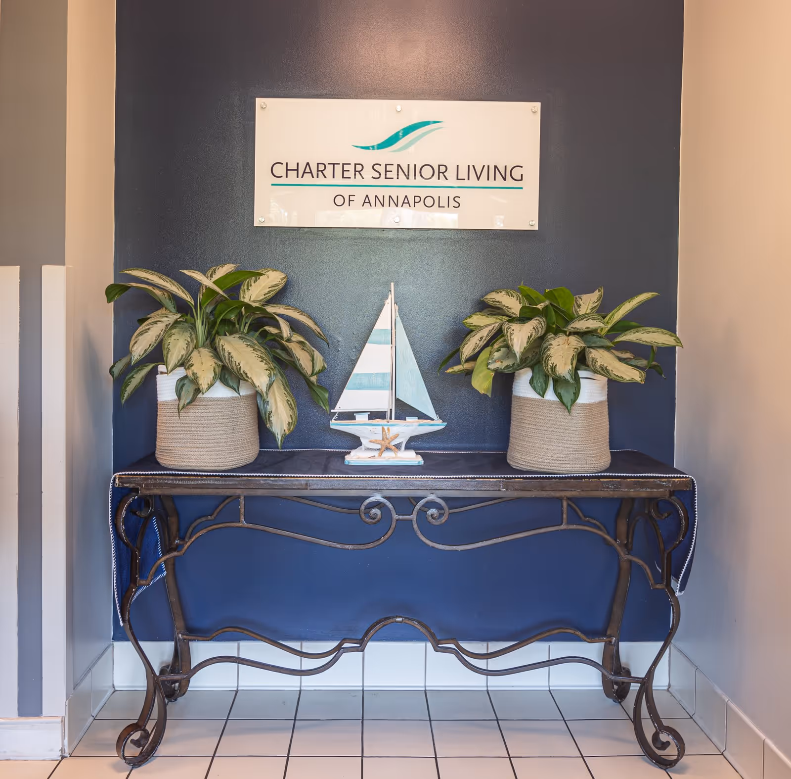 A decorative console table with a dark metal frame and a dark cloth runner is placed against a navy blue wall. On the table, there are two potted plants with green and white leaves on either side of a small decorative sailboat. Above the table, a sign reads 'Charter Senior Living of Annapolis.' The floor is tiled in white.