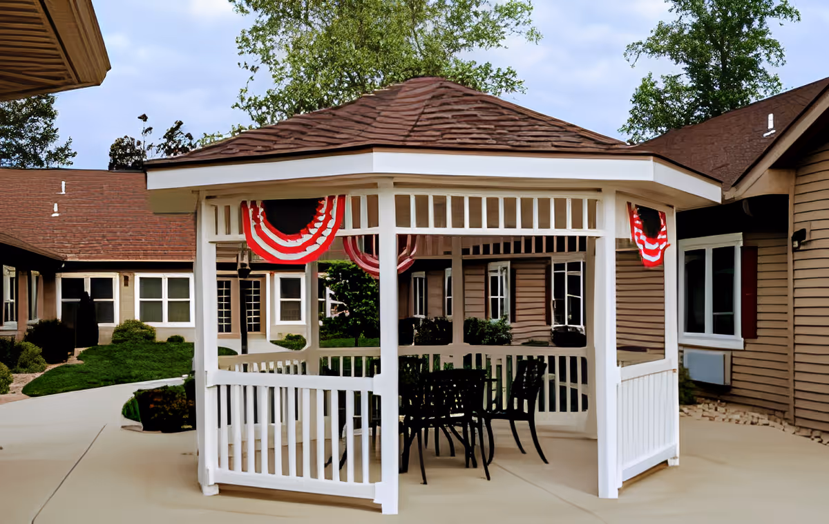 A white gazebo with a brown shingled roof is situated in an outdoor courtyard area of a senior living facility. The gazebo is decorated with red, white, and blue bunting. Inside the gazebo, there is a black metal table with four matching chairs. Surrounding the gazebo are beige buildings with brown roofs and windows, along with some greenery and trees in the background.