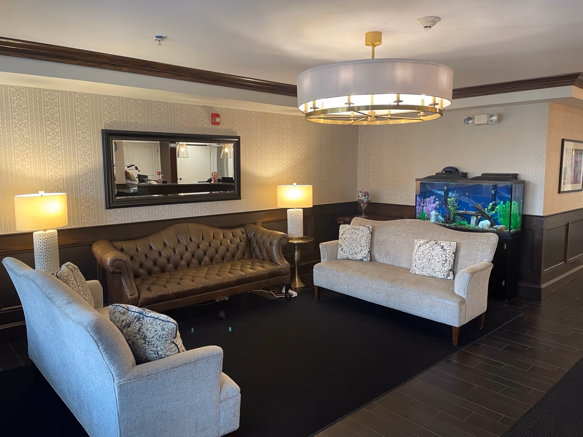 A cozy seating area in a senior living facility with two light gray upholstered sofas and one dark brown tufted leather sofa arranged around a black rug. There are two table lamps with white shades on either side of the leather sofa, a large rectangular mirror on the wall, and a large fish tank filled with colorful aquatic plants and decorations. The room has patterned wallpaper, dark wood paneling, and a modern circular chandelier hanging from the ceiling.