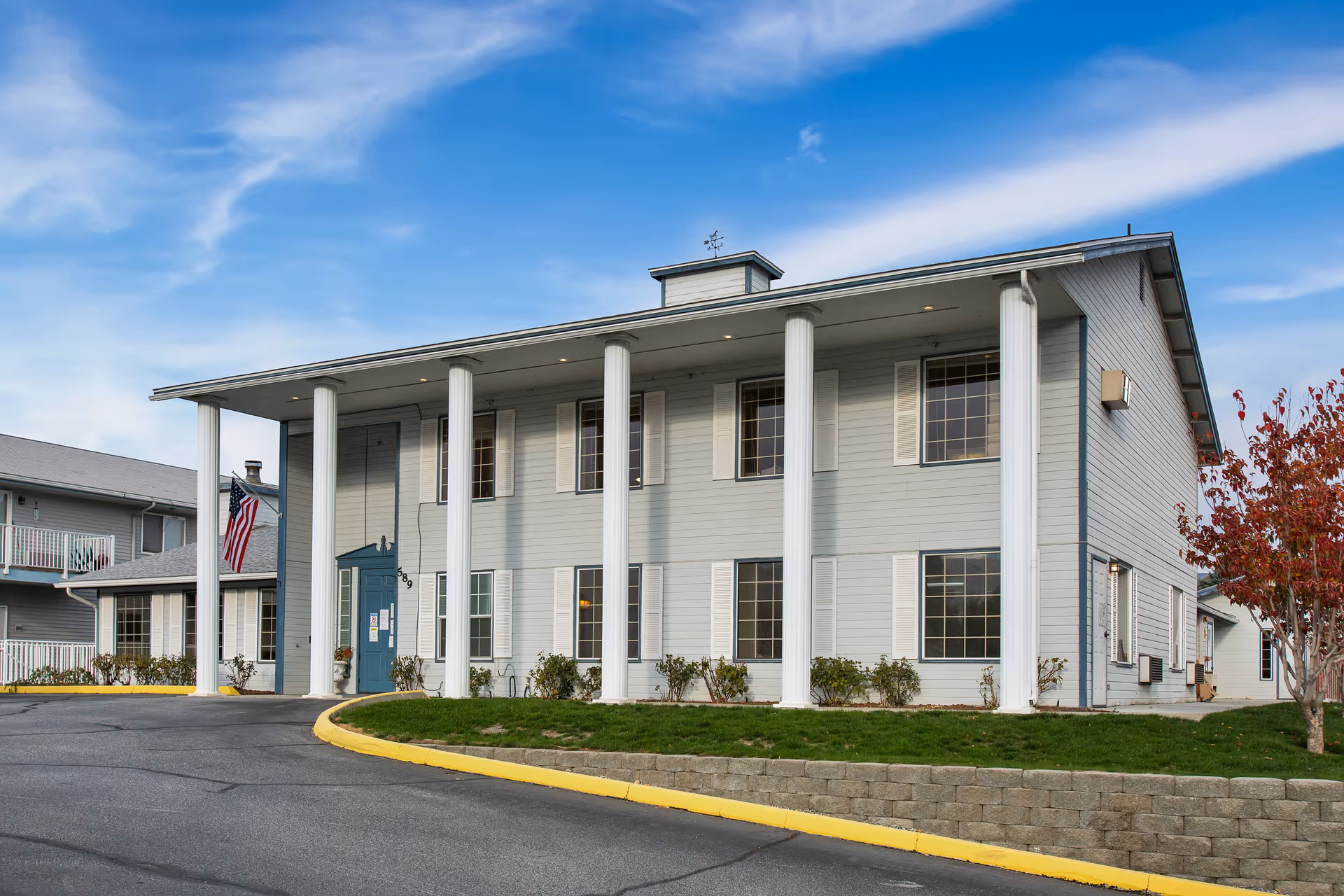 Exterior view of a two-story senior living facility building with white columns, light gray siding, multiple windows with white shutters, a blue entrance door, an American flag, and a small landscaped area with grass and a tree under a partly cloudy blue sky.