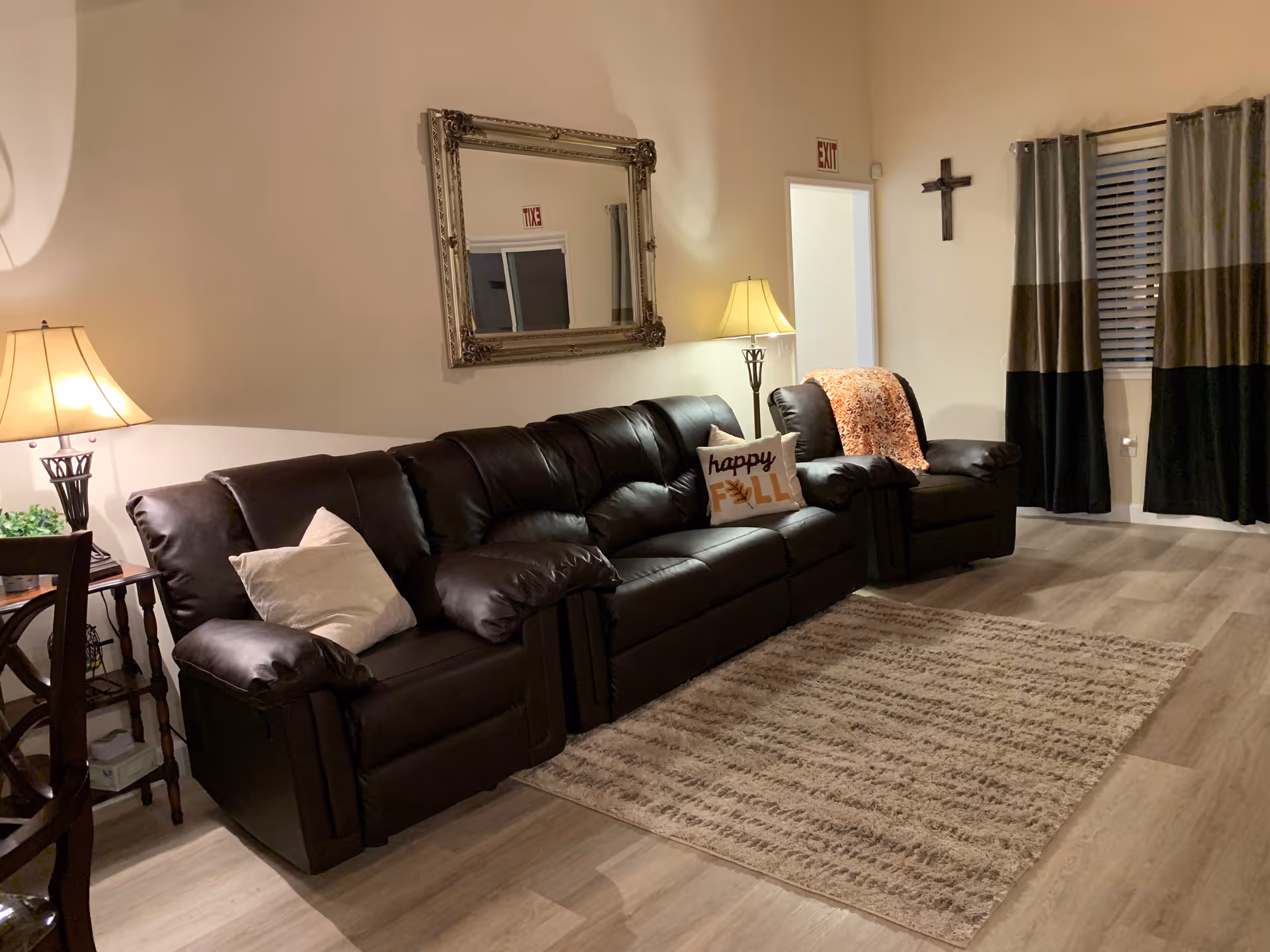 A cozy living room with a dark brown leather sofa and armchair, decorated with pillows including one that says 'happy FALL'. There are two lamps on either side of the sofa, a large ornate mirror on the wall, a cross hanging near a window with dark curtains, and a beige patterned rug on the wooden floor.