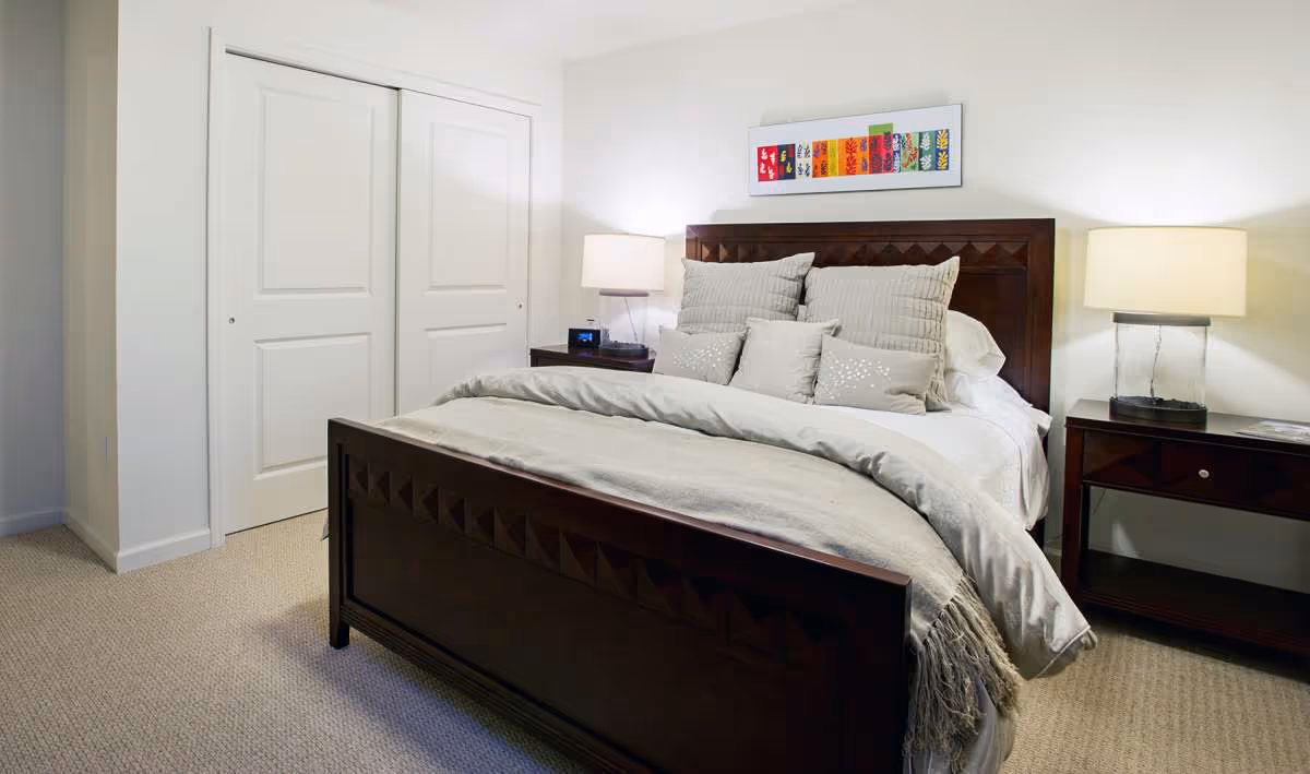 A neatly made bed with a dark wooden headboard and footboard in a bedroom. The bed has multiple pillows and a light gray blanket. There are two matching dark wooden nightstands on either side of the bed, each with a lamp. A colorful framed artwork hangs on the white wall above the bed. A closed white double-door closet is visible on the left side of the room.
