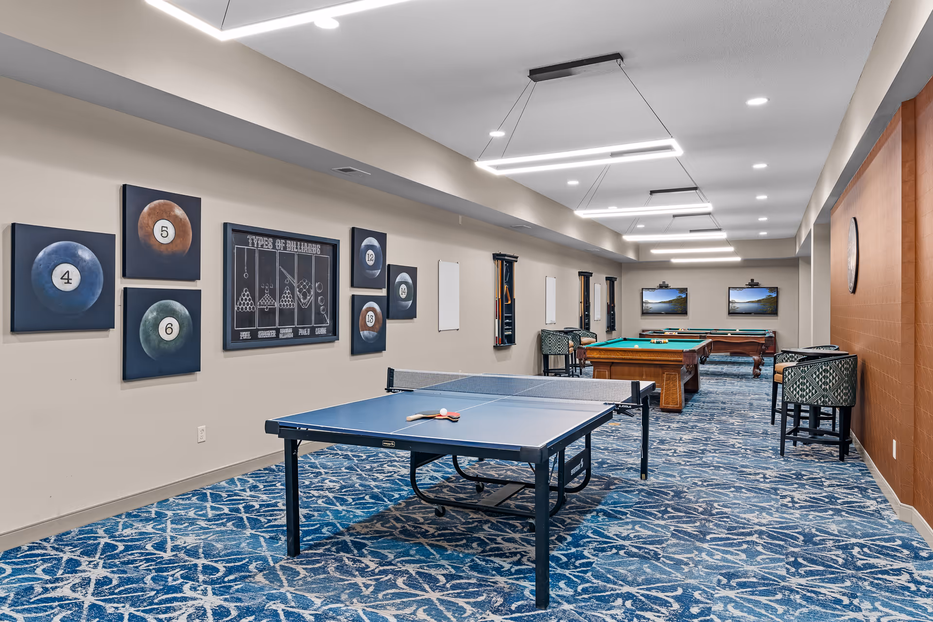 A recreational room with a ping pong table in the foreground and two billiards tables in the background. The walls are decorated with framed pictures of billiard balls and a diagram titled 'Types of Billiards.' The room has patterned blue carpet, modern rectangular ceiling lights, and several chairs along the walls.
