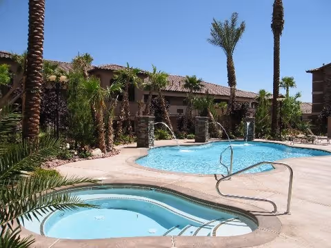 Outdoor swimming pool and hot tub area surrounded by palm trees and landscaping at a senior living facility under a clear blue sky.