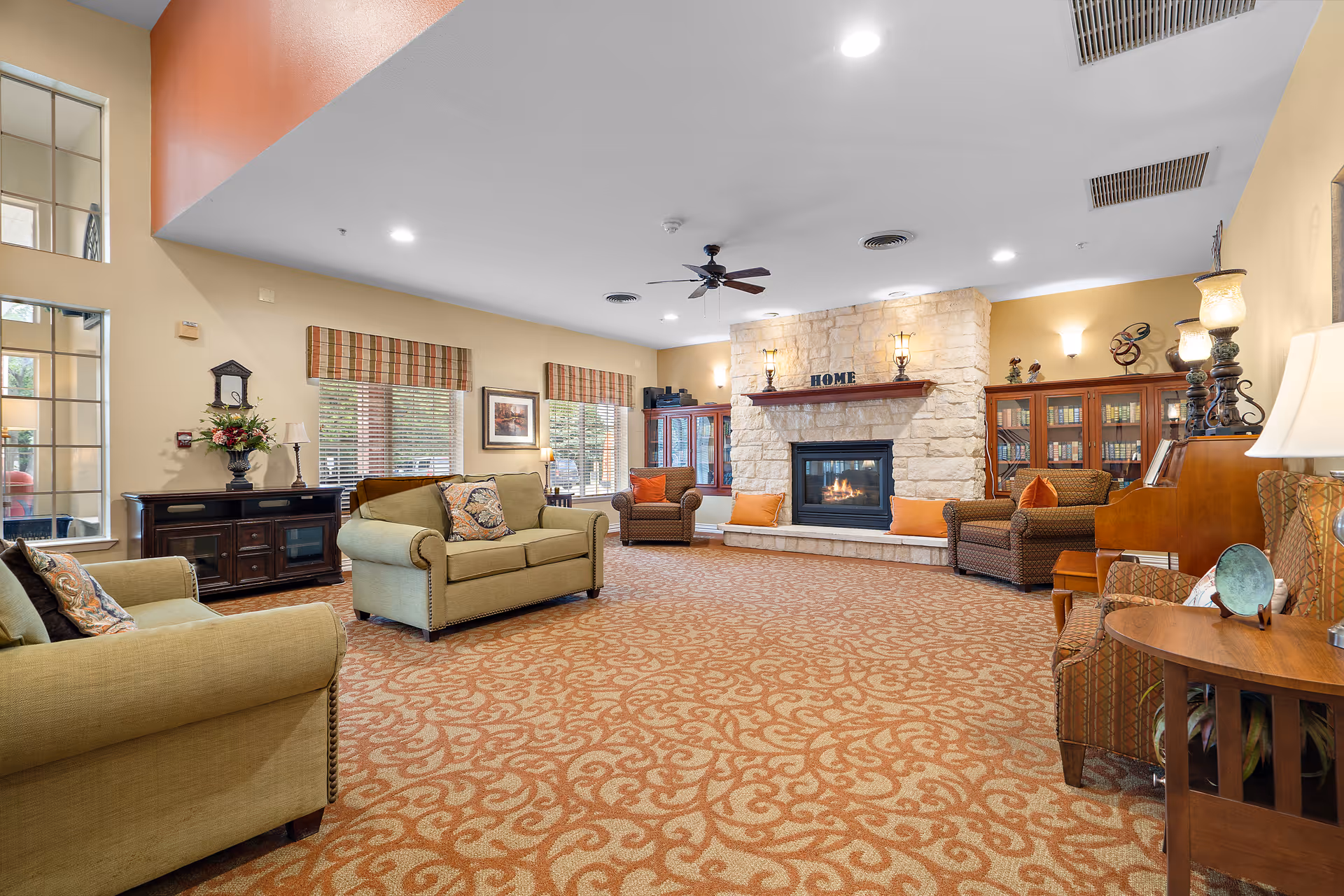 A spacious and warmly decorated living room with patterned carpet, multiple sofas and armchairs, a stone fireplace with the word 'HOME' on the mantle, bookshelves, and large windows with striped valances allowing natural light to enter.