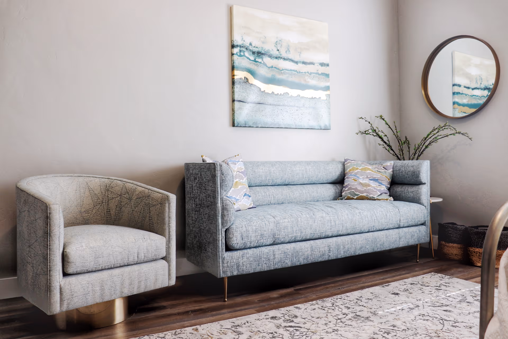 A modern living room with a light gray armchair and a matching light blue-gray sofa with patterned throw pillows. Above the sofa hangs an abstract painting with blue and beige tones. A round mirror is mounted on the adjacent wall, reflecting part of the painting. A small side table with a plant and woven baskets are placed in the corner. The room has wooden flooring and a patterned area rug.