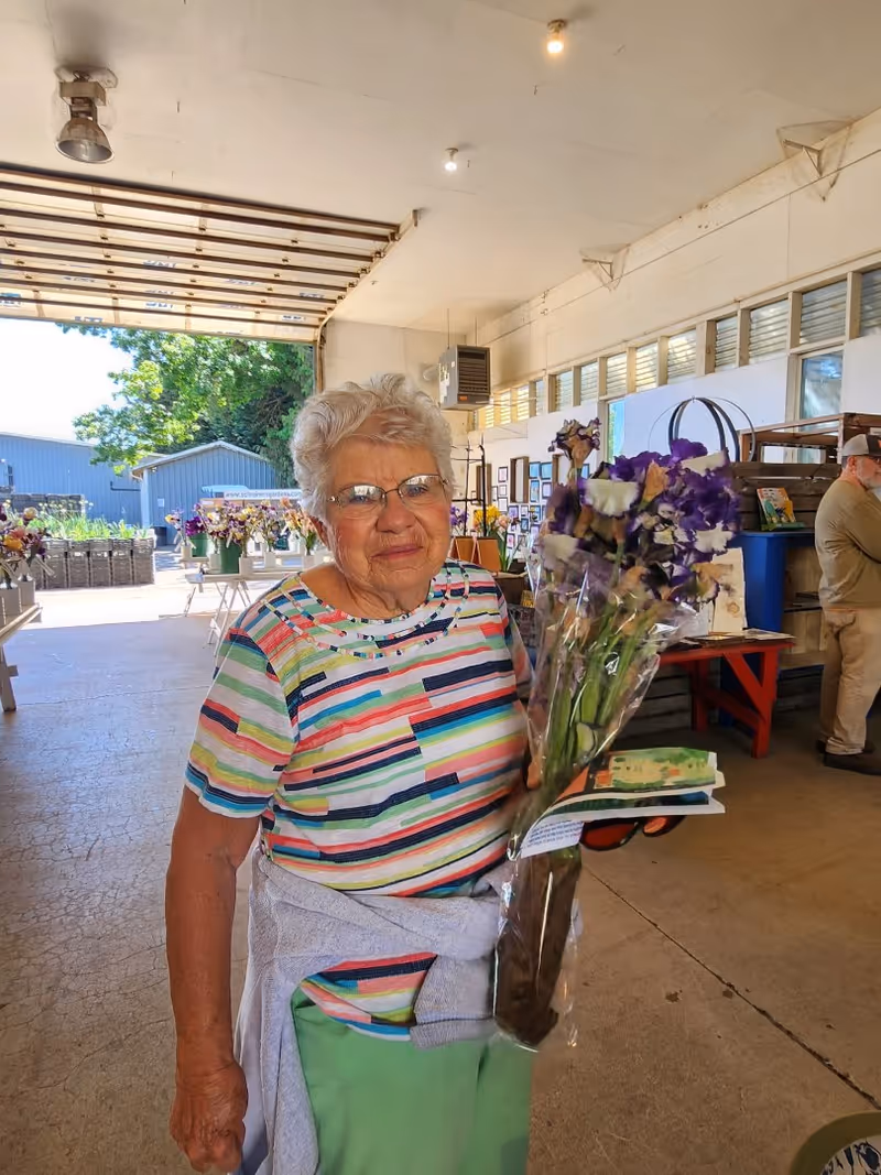 An elderly woman with short white hair and glasses is standing indoors holding a bouquet of purple and white flowers wrapped in plastic. She is wearing a colorful striped shirt and has a light gray sweater tied around her waist. Behind her, there are tables with more flowers and various items, and a man is standing near a wall with windows and framed pictures.