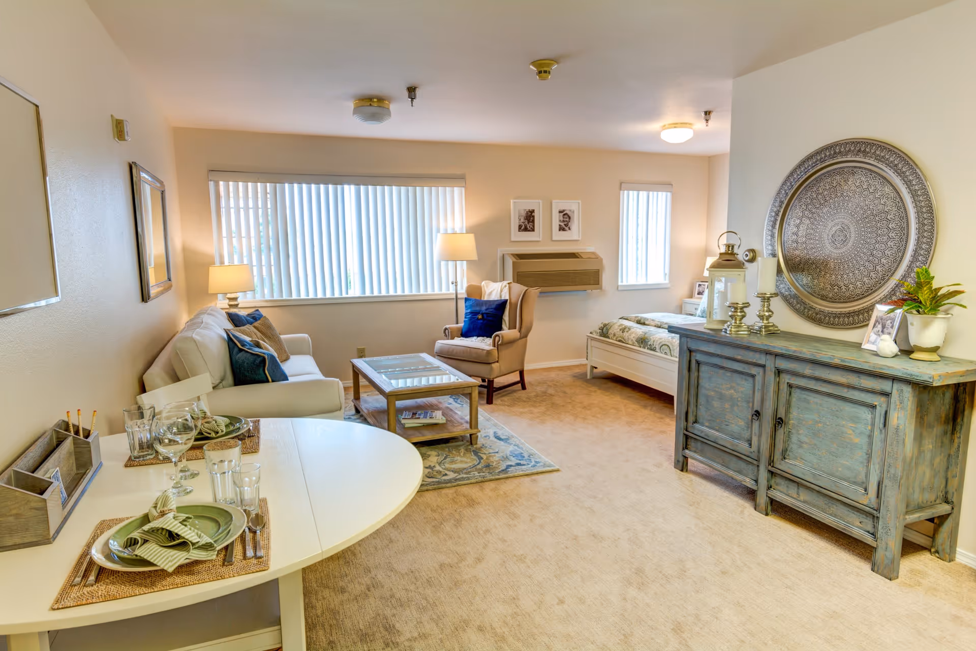 A bright and cozy studio apartment featuring a small dining table set for two with plates, glasses, and napkins. The room includes a white sofa with blue and beige cushions, a beige armchair with a blue pillow, a wooden coffee table on a patterned rug, and a bed with a patterned bedspread. A rustic blue sideboard holds decorative items including candles, a lantern, framed photos, and a potted plant. The walls are light-colored with two windows covered by vertical blinds, and framed pictures hang above a wall-mounted air conditioning unit.