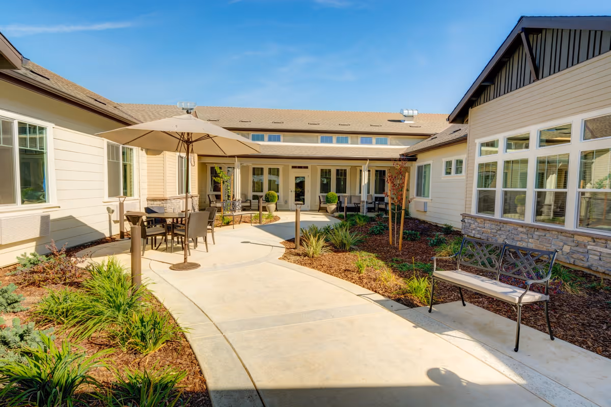 Outdoor courtyard area at The Pavilion at El Dorado Hills featuring a paved walkway, garden beds with plants, patio tables with umbrellas, and benches. The courtyard is surrounded by a beige building with multiple windows under a clear blue sky.