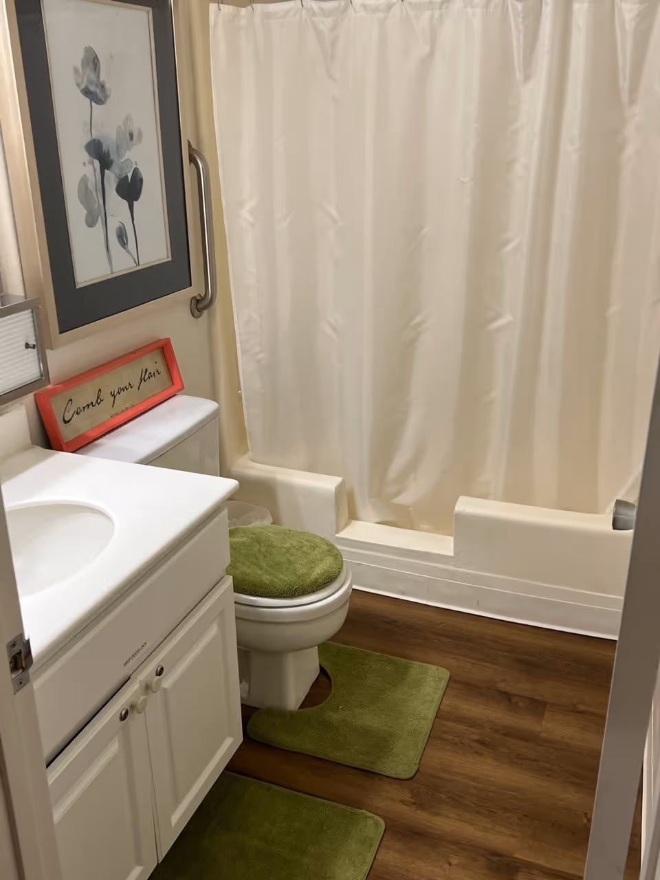 A small bathroom with a white vanity and sink, a toilet with a green cover, green bath mats on a wooden floor, a bathtub with a cream-colored shower curtain, a grab bar on the wall, and framed artwork and a sign that says 'Comb your hair'.