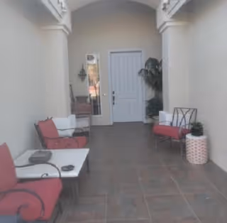 Covered outdoor patio area with tiled floor, featuring red cushioned chairs, a small white table, a potted plant, and a white door at the end of the space.