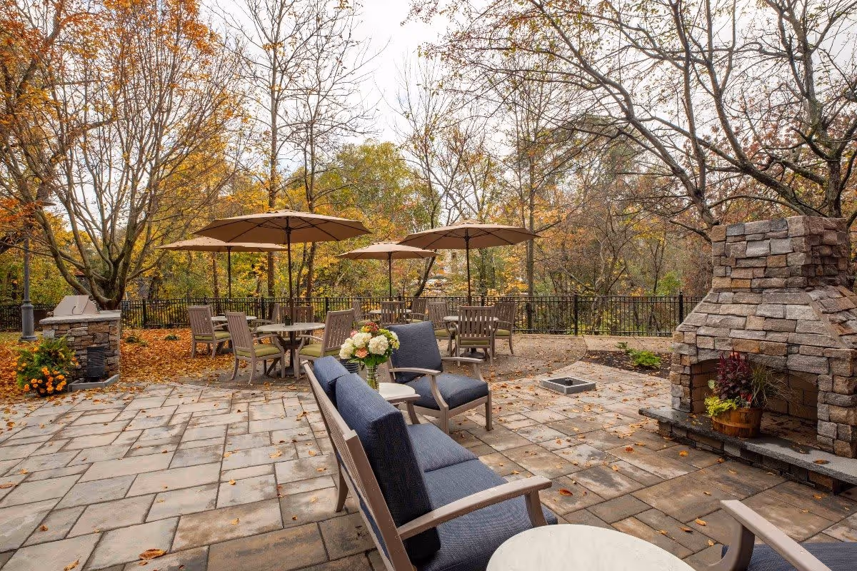 Outdoor patio area with stone flooring, several tables with umbrellas, cushioned chairs, and a stone fireplace surrounded by autumn trees with fallen leaves.
