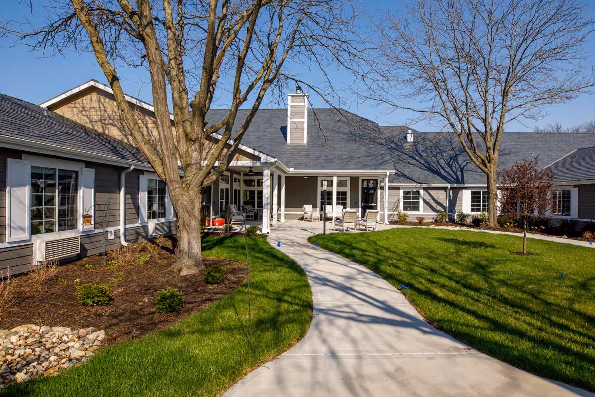 Single-story assisted living building with a paved walkway, green lawn, mature trees, and outdoor seating at the main entrance.
