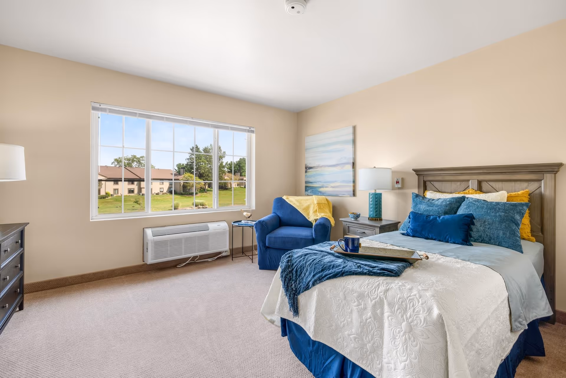 A bright and cozy bedroom with a large window showing a view of green lawns and buildings outside. The room features a bed with blue and yellow pillows and a white textured bedspread, a blue armchair with a yellow throw, a nightstand with a lamp and decorative items, a dresser with a lamp, and a piece of artwork on the wall.