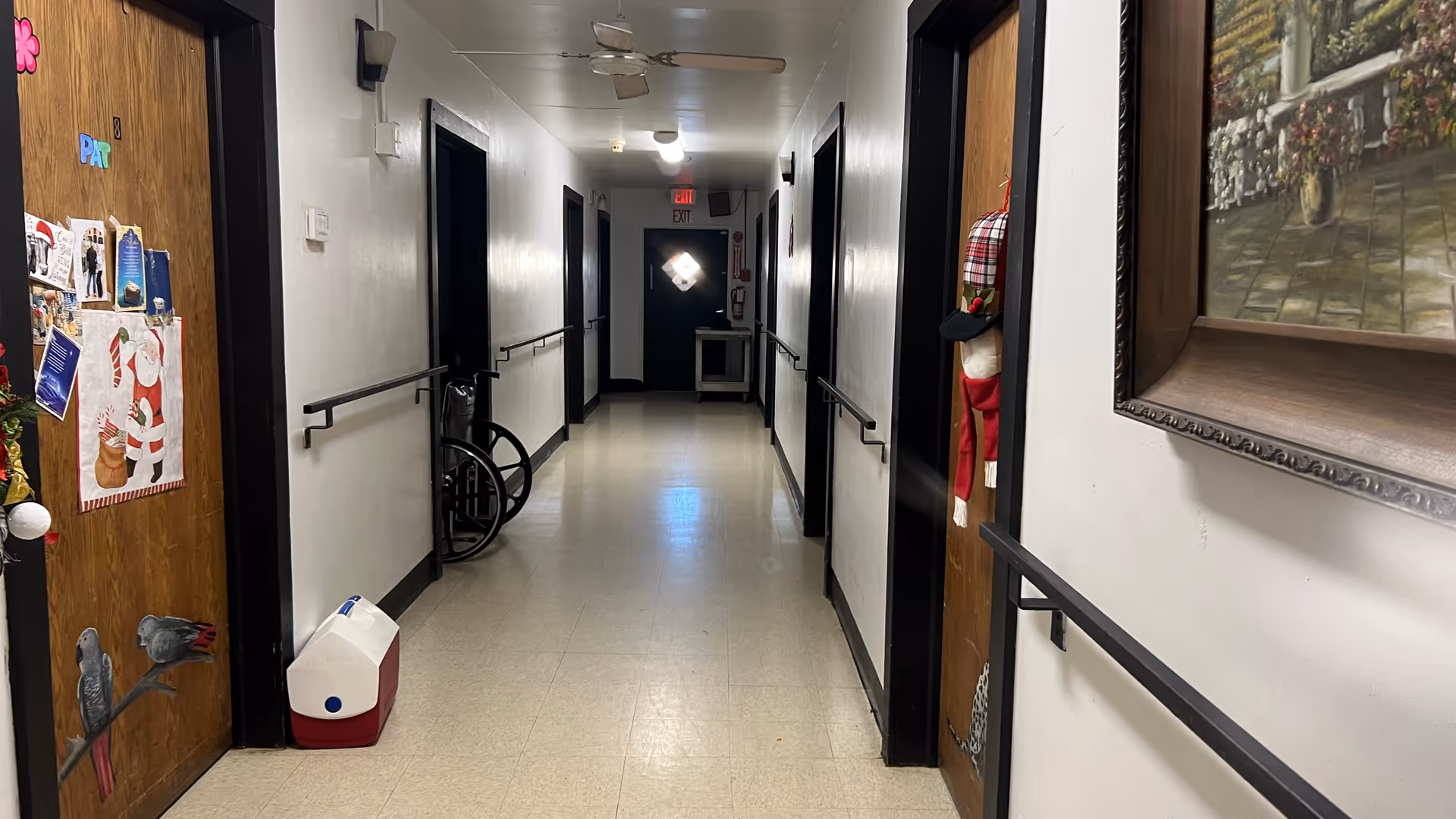 Interior hallway of a senior living facility with handrails, decorated doors, a wheelchair, and a cooler along the corridor.