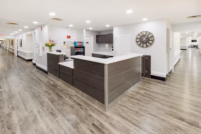 Reception desk and nursing station in a bright senior living hallway with wood-look floors and a large wall clock.
