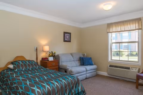 A senior living facility bedroom with a bed covered in a brown and teal patterned comforter, a wooden nightstand with a lamp and flowers, a gray patterned loveseat with a blue pillow, a window with beige curtains, and a wall-mounted air conditioning unit below the window.
