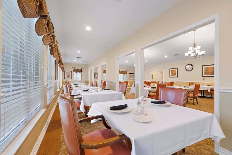 A bright dining room with multiple tables covered in white tablecloths, each set with plates, napkins, and silverware. The room has large windows with blinds and brown valances, and red cushioned chairs around the tables. The walls are light-colored with framed artwork and a clock visible in the background.