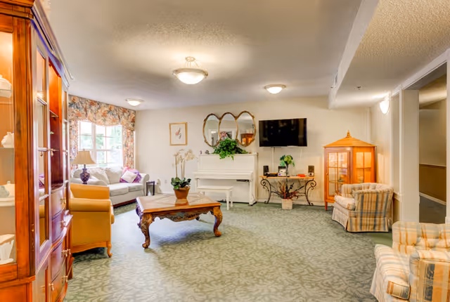 A cozy living room in a senior living facility featuring a white piano with a decorative mirror above it, a flat-screen TV mounted on the wall, a wooden coffee table with a potted plant, a glass display cabinet, and several upholstered chairs and sofas arranged around the room. The room has patterned carpet, floral curtains on the window, and soft ceiling lighting.