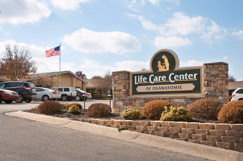 Stone sign for Life Care Center of Osawatomie surrounded by landscaping with bushes, a parking lot with cars, and an American flag on a flagpole in the background under a partly cloudy sky.