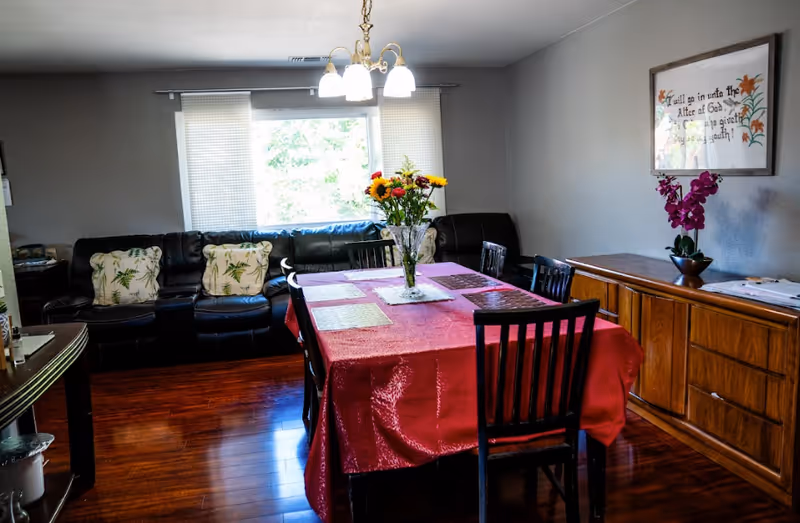 A dining room with a red-cloth table, chairs, flowers, and a sideboard by windows