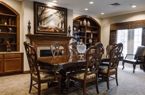 A traditional dining room with a polished wooden dining table surrounded by six ornate chairs with cushioned seats. Behind the table is a fireplace with a large framed painting of a tree-lined path above it. On either side of the fireplace are built-in wooden shelves displaying decorative items. To the right, there is a window with patterned curtains and a dark upholstered armchair nearby. The room is warmly lit with recessed ceiling lights.