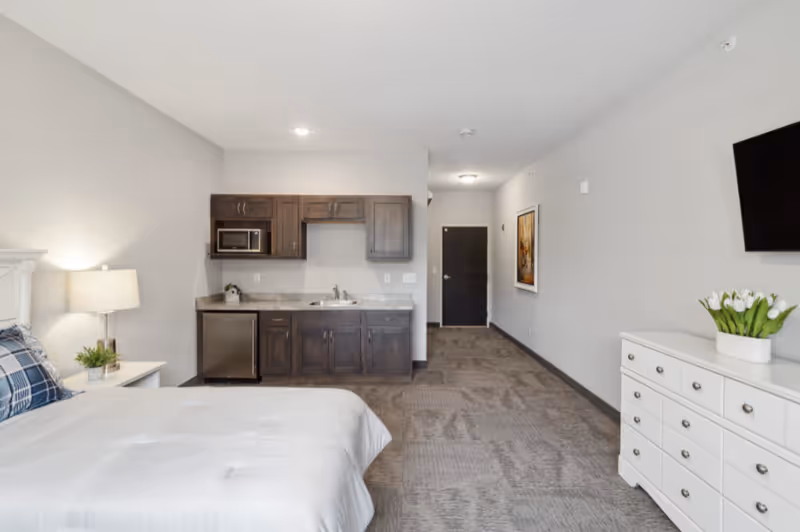 Studio-style senior living bedroom with a bed, nightstand, kitchenette, dresser and wall-mounted TV in a neutral-toned interior.