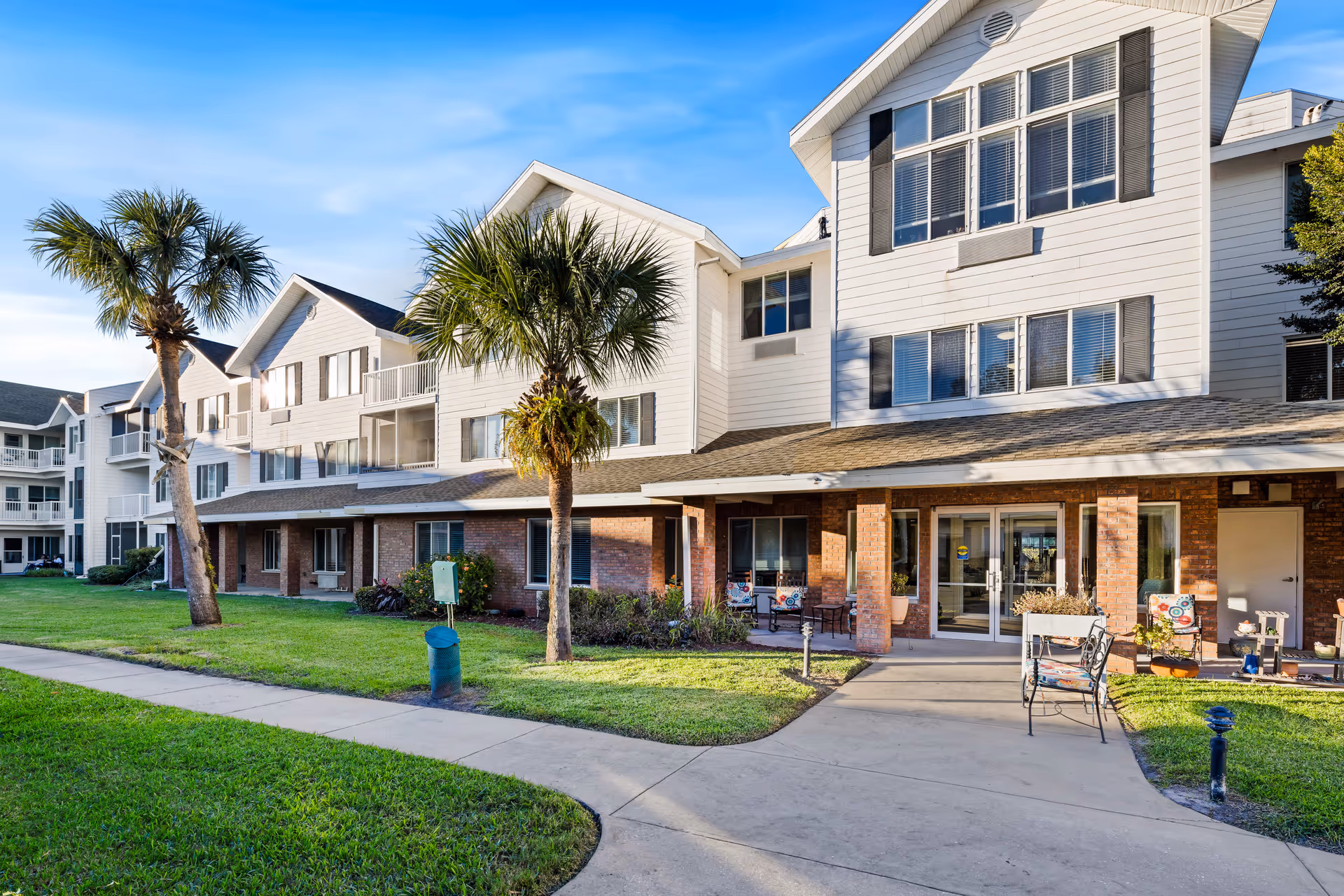 Exterior view of a senior living community building with white siding and brick accents, featuring multiple windows and balconies. There are palm trees and a well-maintained lawn with a concrete walkway leading to the entrance. Outdoor seating is visible near the entrance under a covered porch.