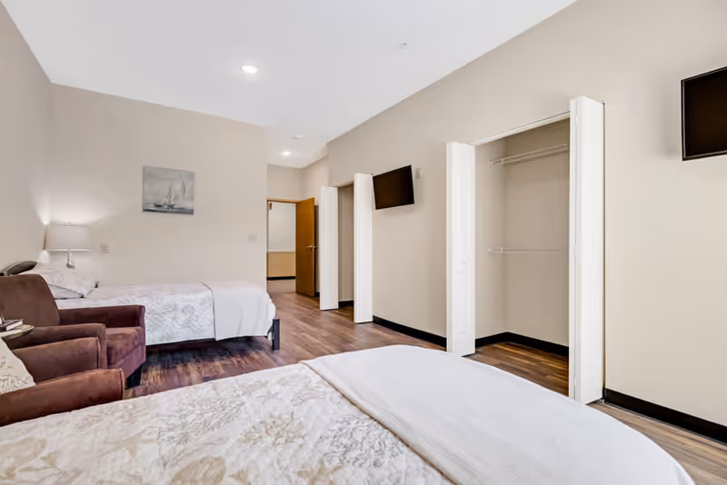 A bright and clean bedroom in an assisted living facility featuring two beds with white and beige bedding, a brown armchair, an open closet with white bi-fold doors, a wall-mounted flat screen TV, wooden flooring, and a painting of a sailboat on the wall.