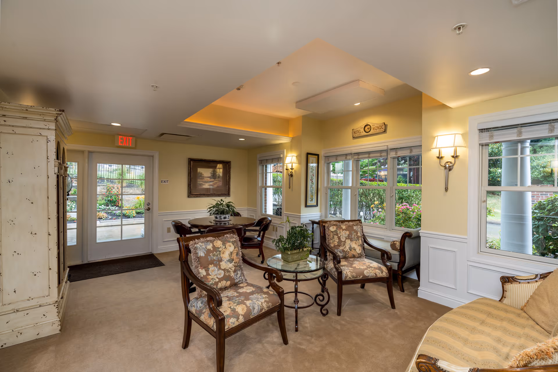 A bright lounge area with floral-upholstered chairs around a glass-top table, large windows looking out to a garden, and an entrance door with an EXIT sign.