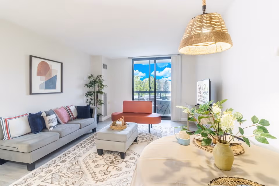 Bright and modern living room with a gray sofa adorned with colorful pillows, a red bench near a sliding glass door leading to a balcony, a patterned area rug, a small ottoman with a basket on top, a round table with a vase of flowers, and a hanging woven light fixture.