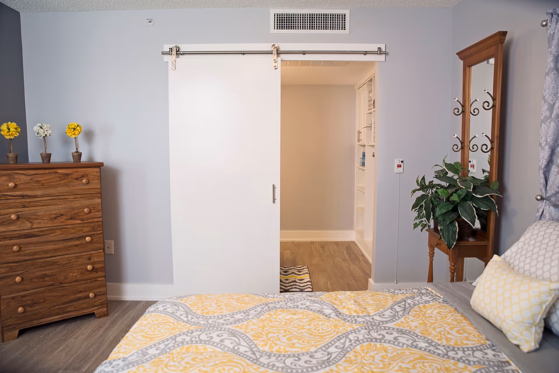 A bedroom with a bed covered in a yellow and gray patterned comforter and pillows. To the left is a wooden dresser with three small potted flowers on top. A white sliding barn door is partially open, revealing a hallway with built-in shelves. On the right side of the room is a wooden mirror stand with a green potted plant in front of it.