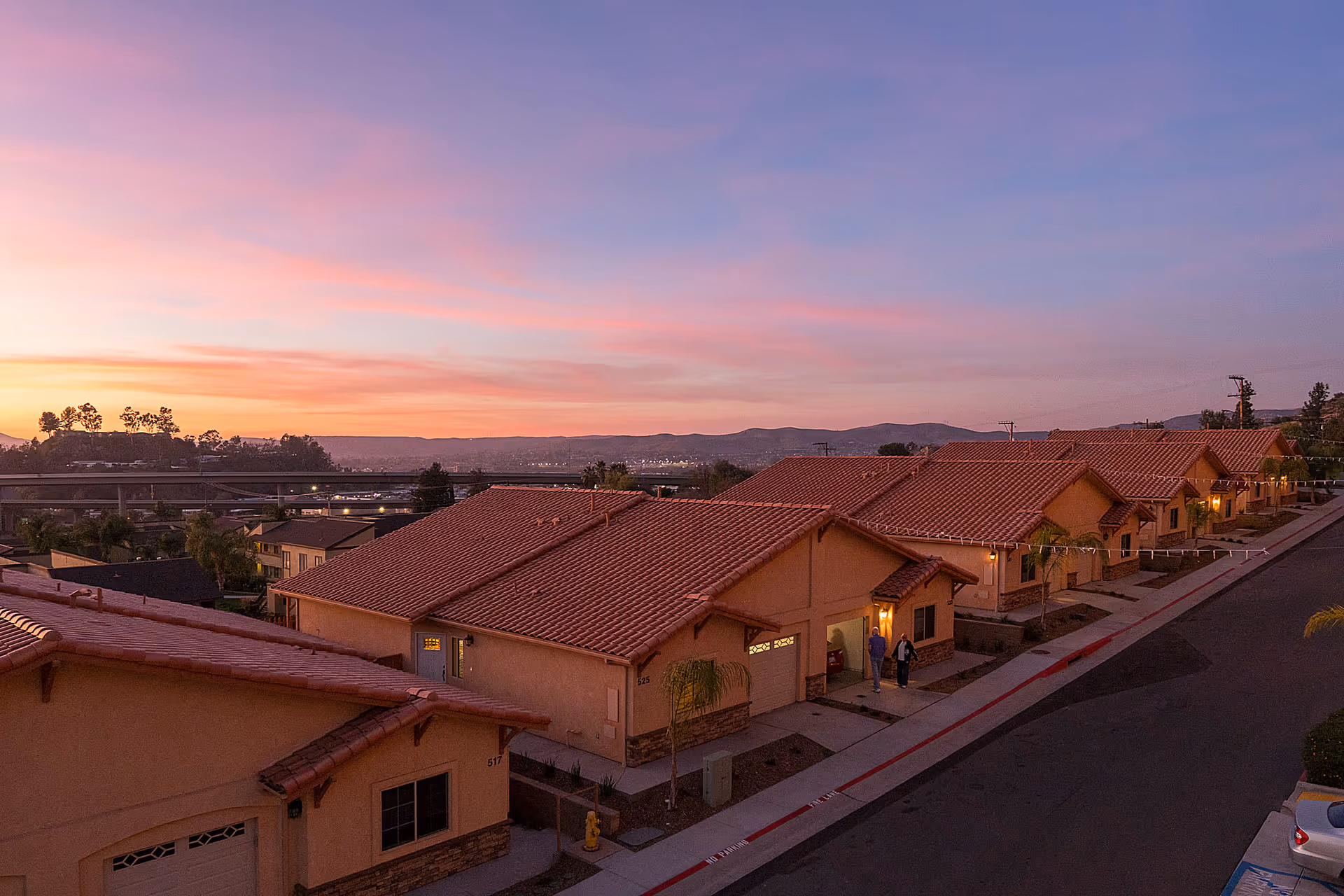Row of single-story tile-roof senior living cottages along a street at sunset with distant hills.