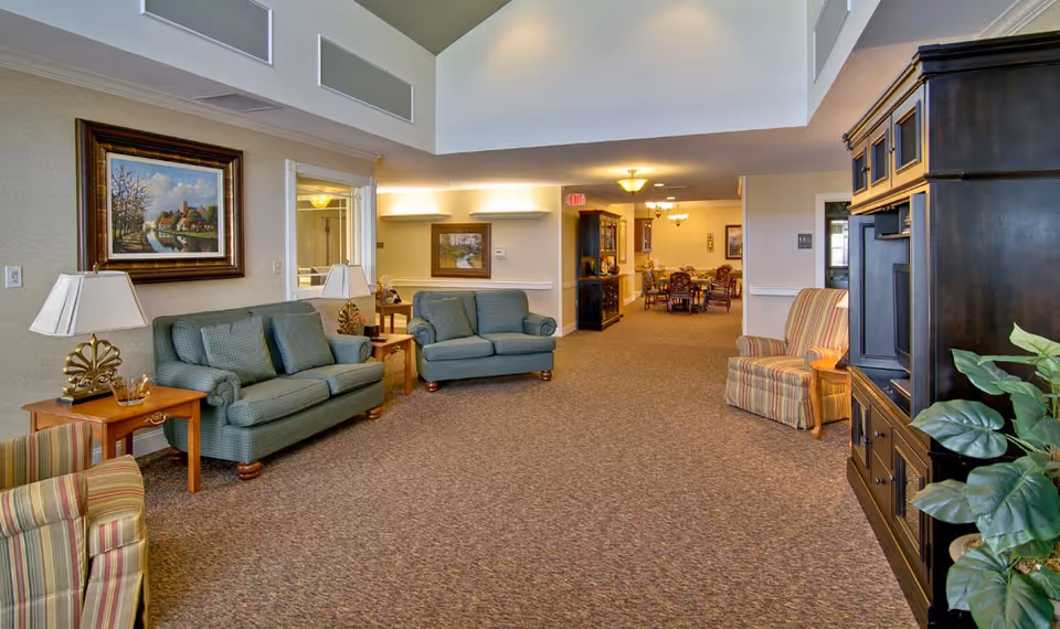 A spacious senior living common room with sofas, armchairs, side tables, and a TV cabinet opening onto a dining area.