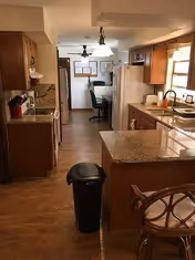 Interior view of a kitchen with wooden cabinets, granite countertops, a refrigerator, microwave, and a trash bin in the center. There is a small desk area with a chair and a ceiling fan in the background.