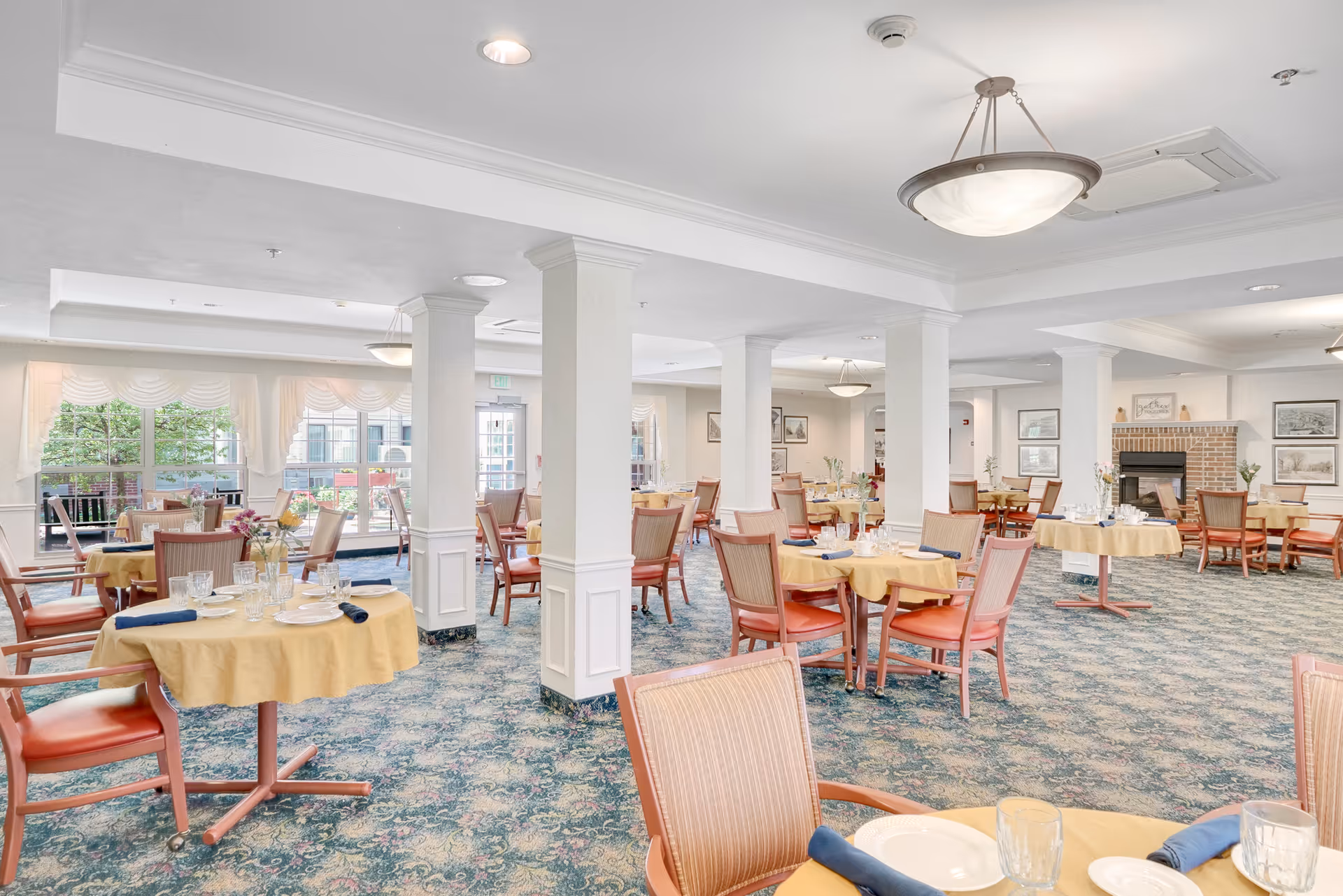 A spacious dining room with multiple round tables covered with yellow tablecloths, each set with plates, glasses, and blue napkins. The room features large windows letting in natural light, white columns, patterned carpet, and framed artwork on the walls. There is a brick fireplace in the background.