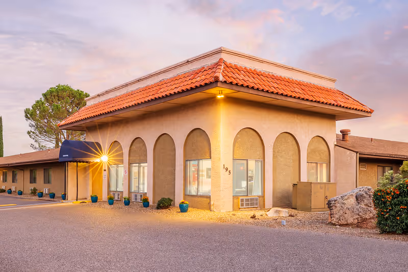 Exterior view of a single-story building with a beige stucco finish and a red tile roof at sunset. The building has several arched windows and a blue awning over the entrance. There are potted plants along the side and a large rock and shrubbery near the corner.