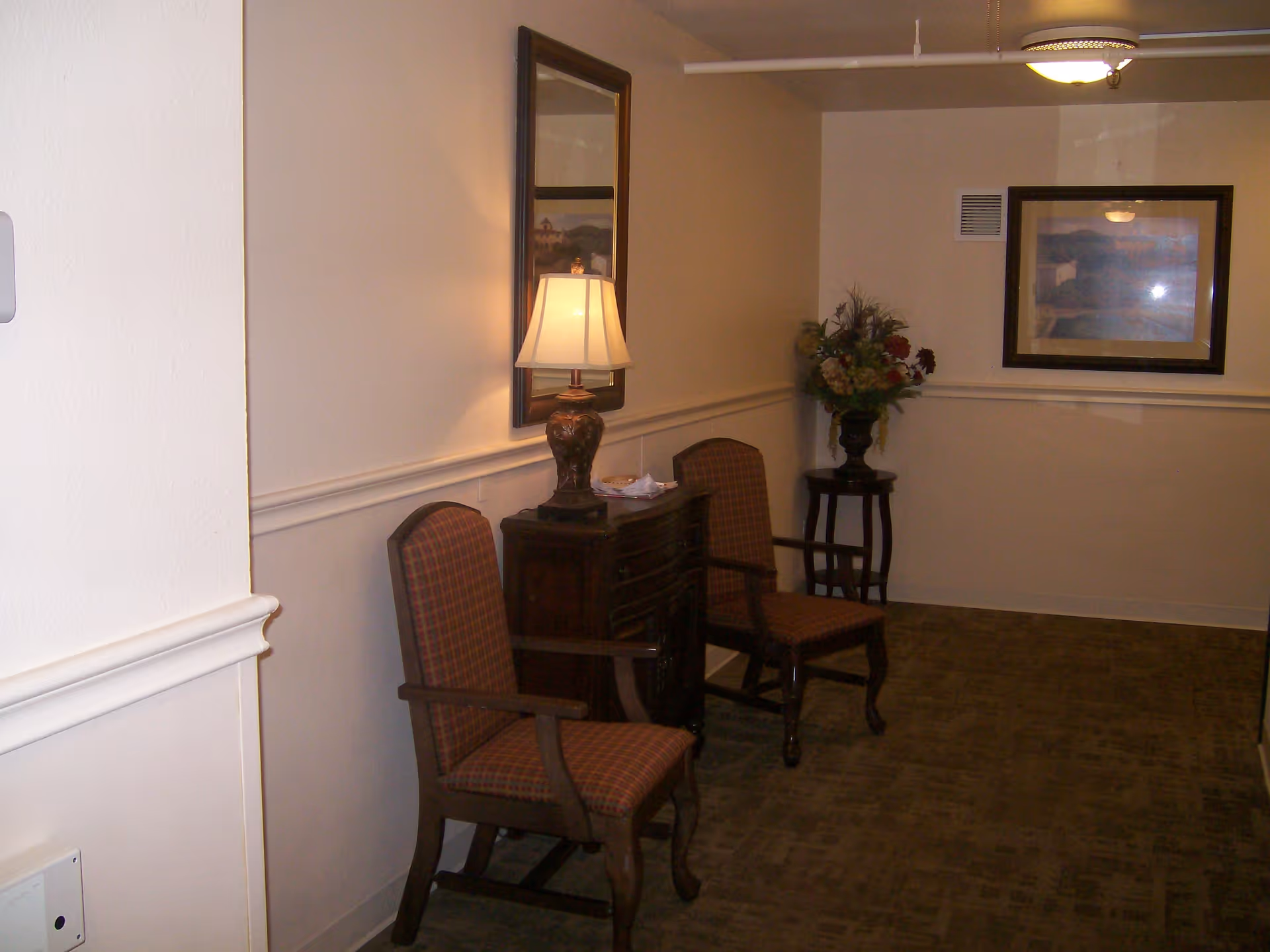 A hallway area with two wooden armchairs with patterned upholstery, a small wooden cabinet with a table lamp on top, a framed mirror above the cabinet, a small round table with a floral arrangement, and a framed picture on the far wall.