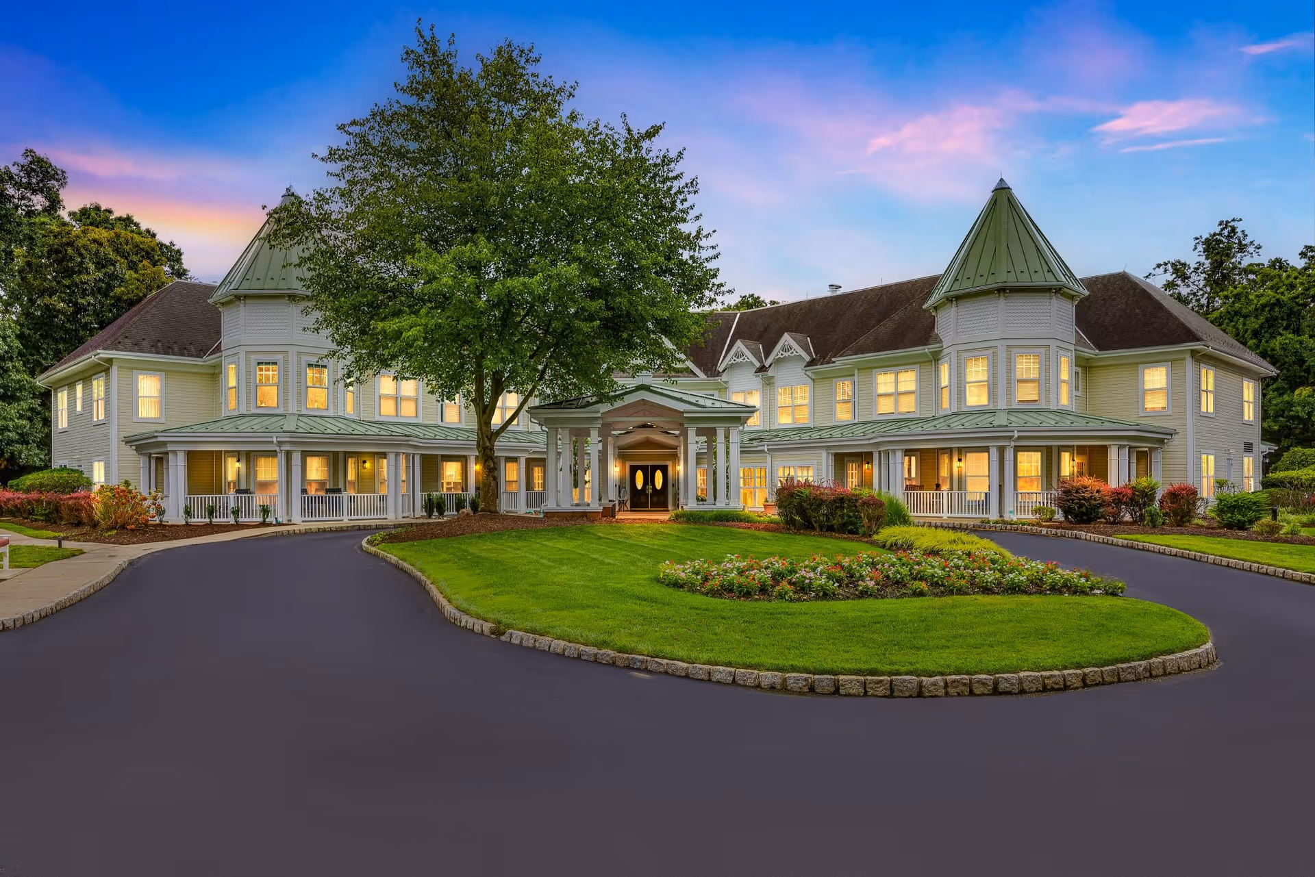 Exterior view of a large, elegant senior living facility building at sunset with a circular driveway, well-maintained lawn, flower beds, and a large tree in front. The building has two prominent turret-like structures with green roofs and many lit windows.