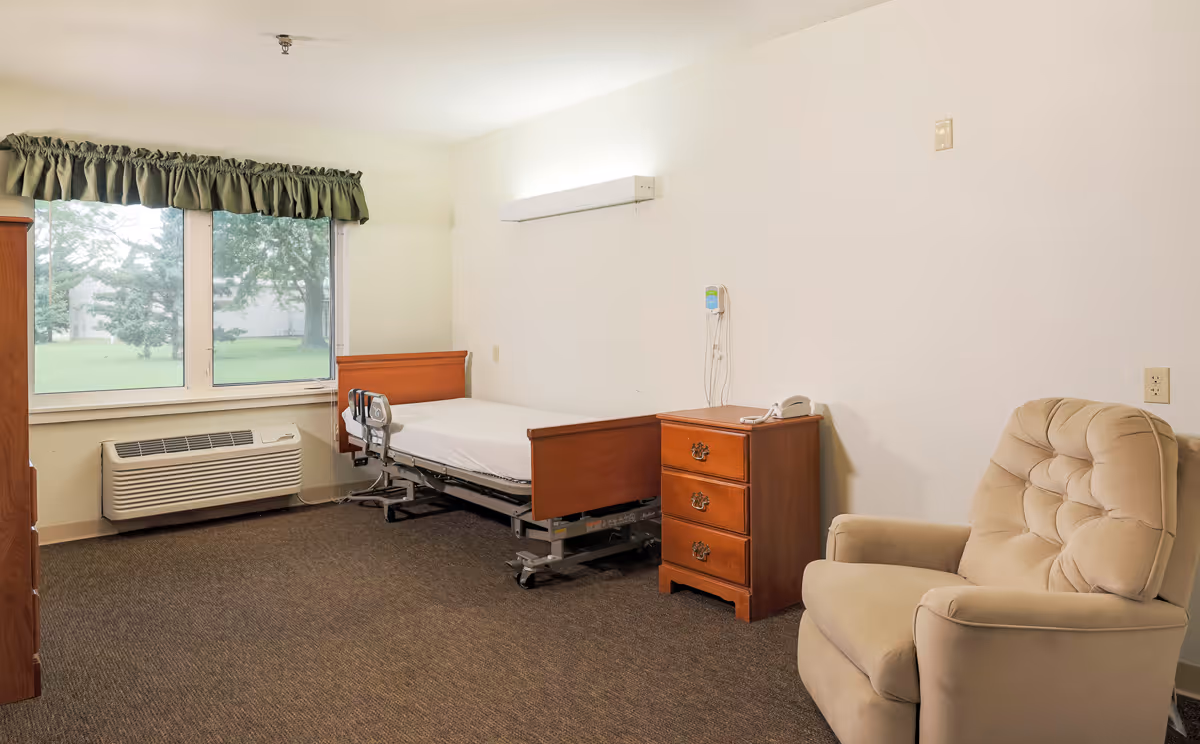 A simple, clean room with a hospital-style bed, a wooden nightstand with a telephone, a beige cushioned armchair, a large window with green curtains, and a wall-mounted air conditioning unit below the window.